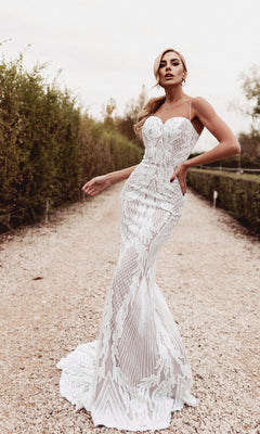 A woman stands on a gravel path outdoors, surrounded by shrubs, wearing the Long White Mermaid Wedding Dress: Tina Holly BB050 with intricate patterns.