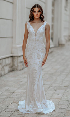 A woman models the Classic Long White Formal Dress: Tina Holly BB052, featuring a fitted silhouette, deep V-neckline, and intricate lace patterns, standing on a stone walkway by a light-colored building.