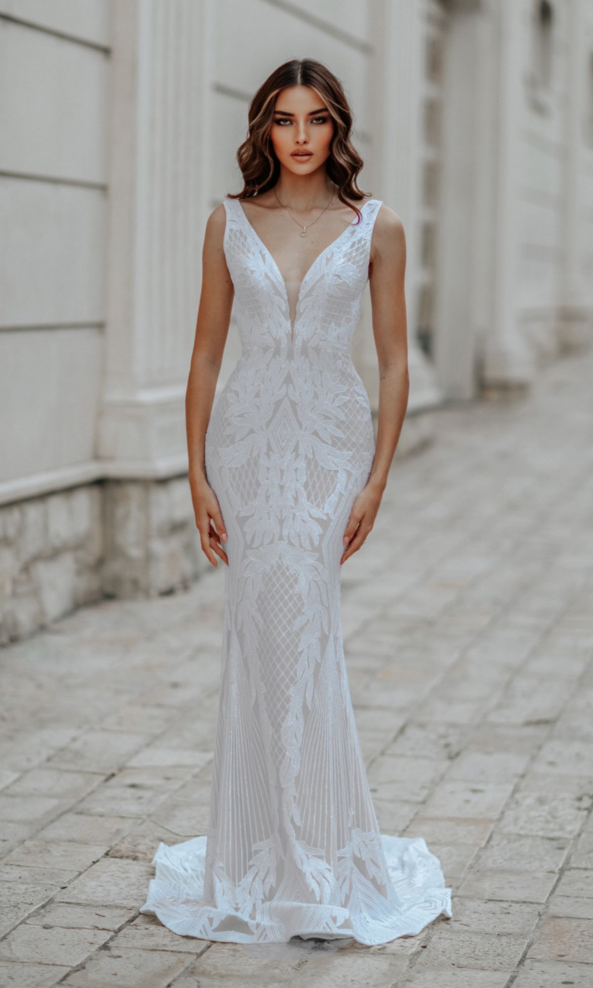 A woman stands outdoors in the Classic Long White Formal Dress: Tina Holly BB052, a sleeveless, form-fitting gown with lace details, deep V-neck, and subtle train, set against a light stone wall background.