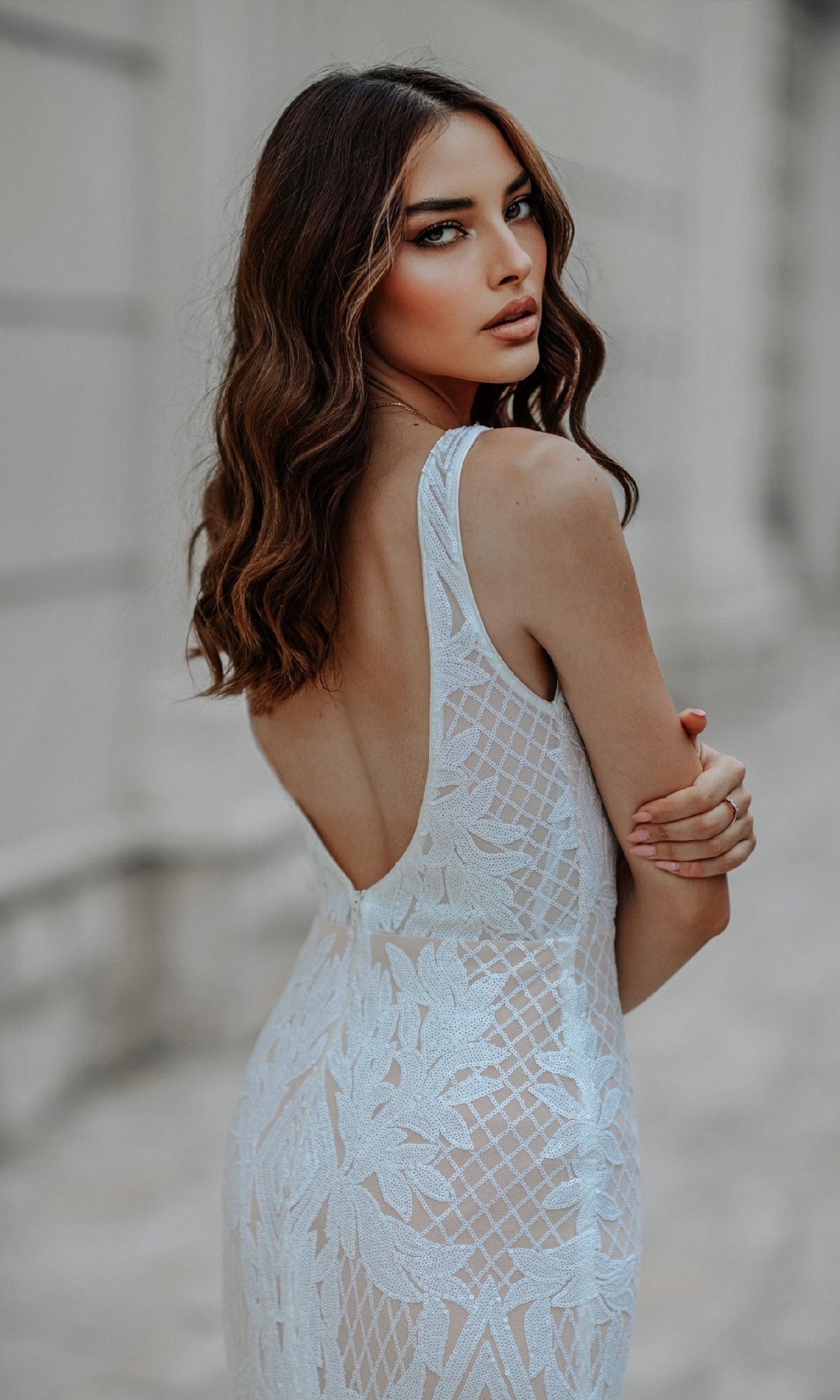 A woman with wavy brown hair wears the Classic Long White Formal Dress: Tina Holly BB052, a sleeveless, open-back gown, standing outdoors and looking over her shoulder at the camera.