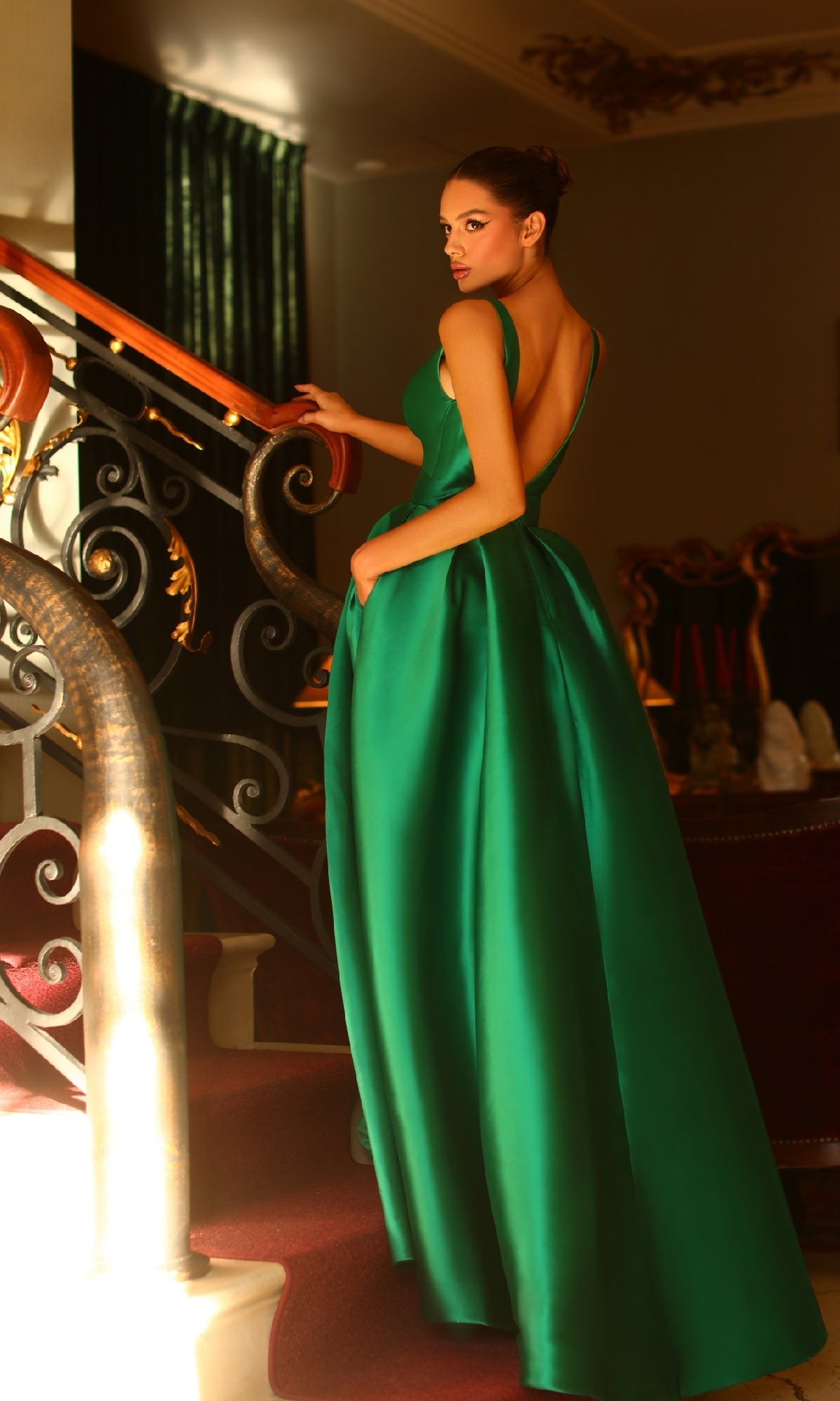 A woman in the Belted Long Prom Ball Gown: Tina Holly BB209, a sleeveless, floor-length green dress, stands on a staircase and looks back over her shoulder in an elegant, warmly lit interior.