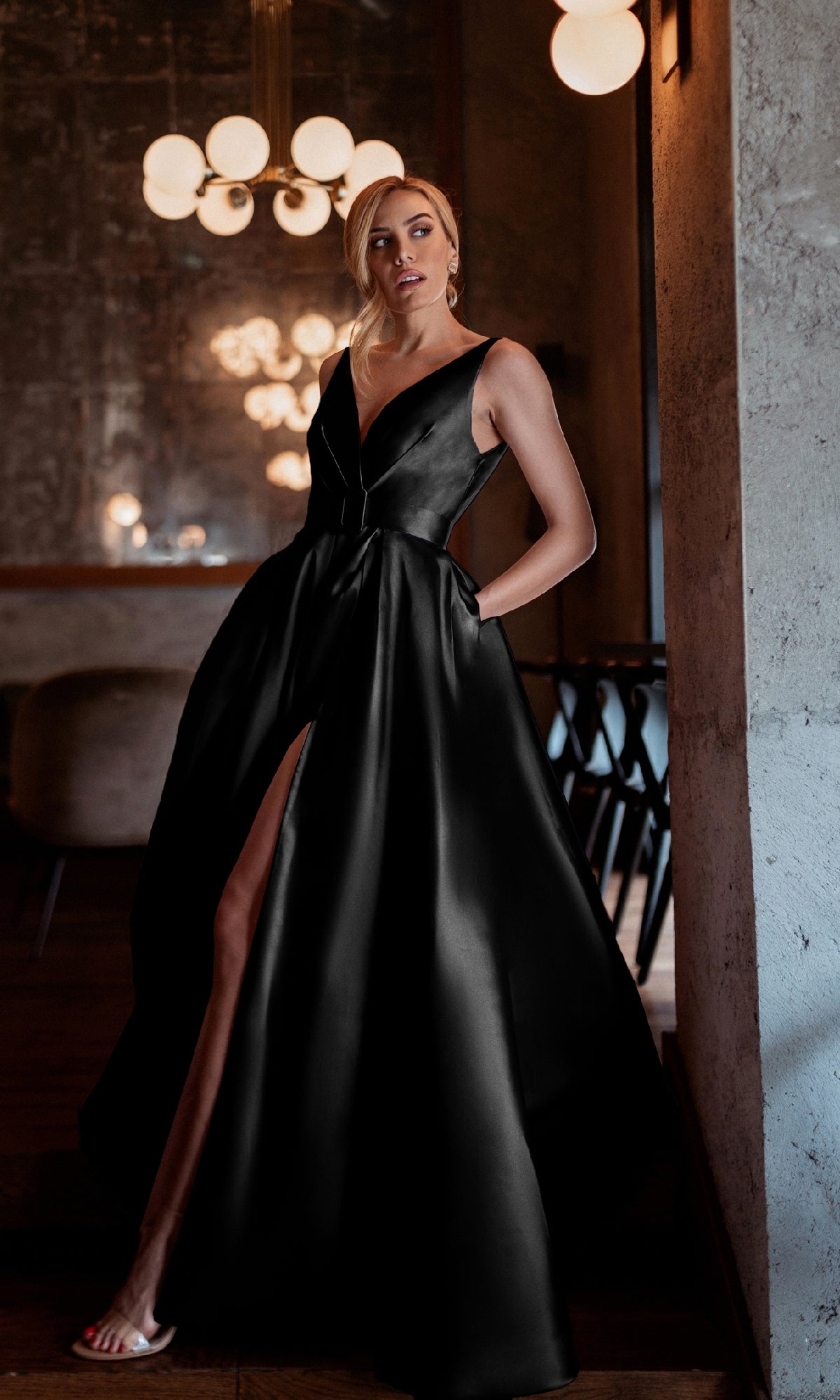 A woman poses indoors with one hand in her pocket, wearing the Tina Holly BB209 Belted Long Prom Ball Gown—a sleeveless, floor-length black dress featuring a deep V-neck and a high slit.