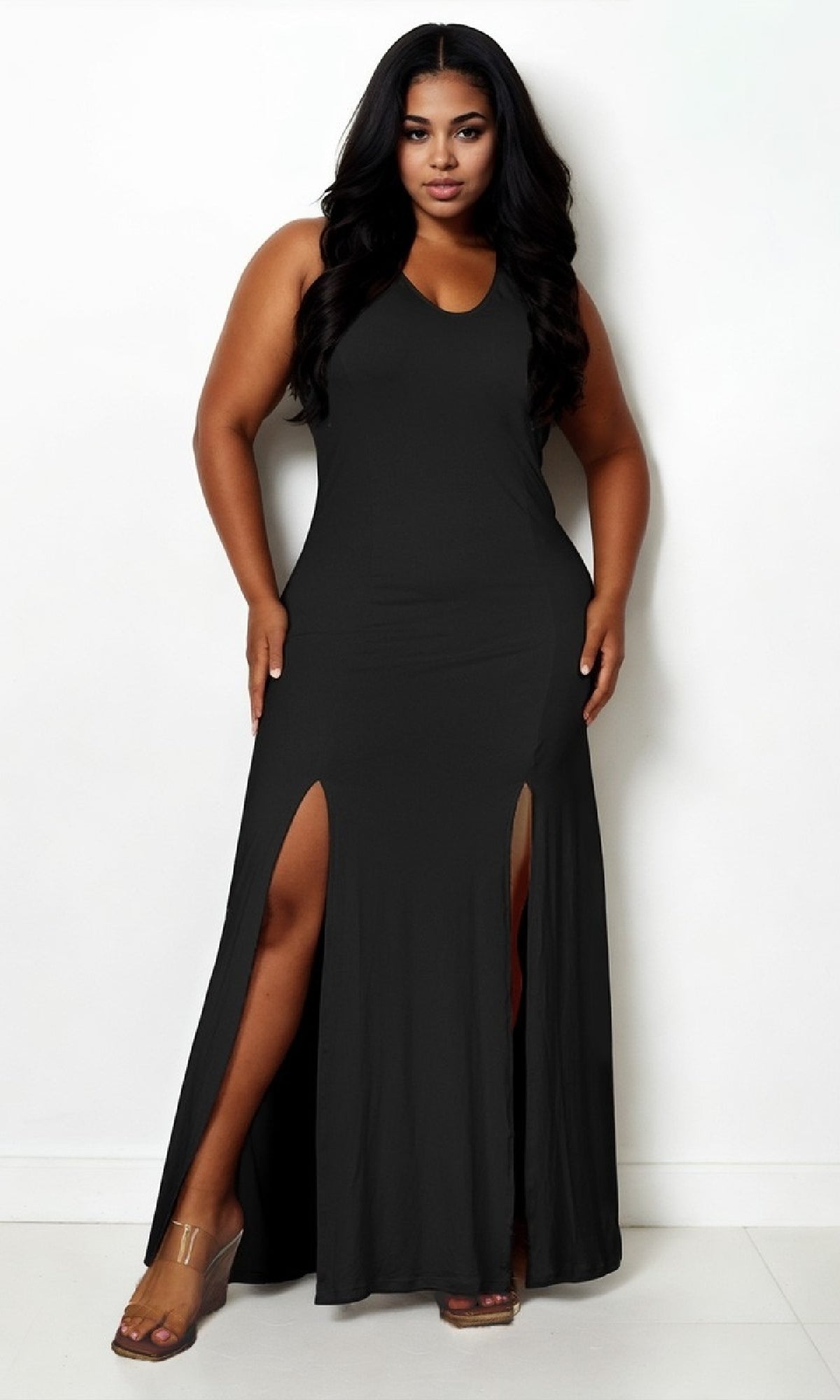 A woman models the black Sleeveless Double-Slit Casual Plus-Size Maxi Dress against a white wall, pairing this v-neck maxi dress with clear high-heeled sandals.