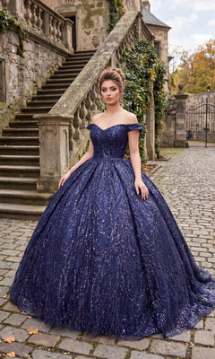 A woman stands on a cobblestone path in front of stone stairs, wearing the Quinceanera Dress BT9013 By Chicas in navy, featuring glittering embellishments and an elegant off-the-shoulder neckline.