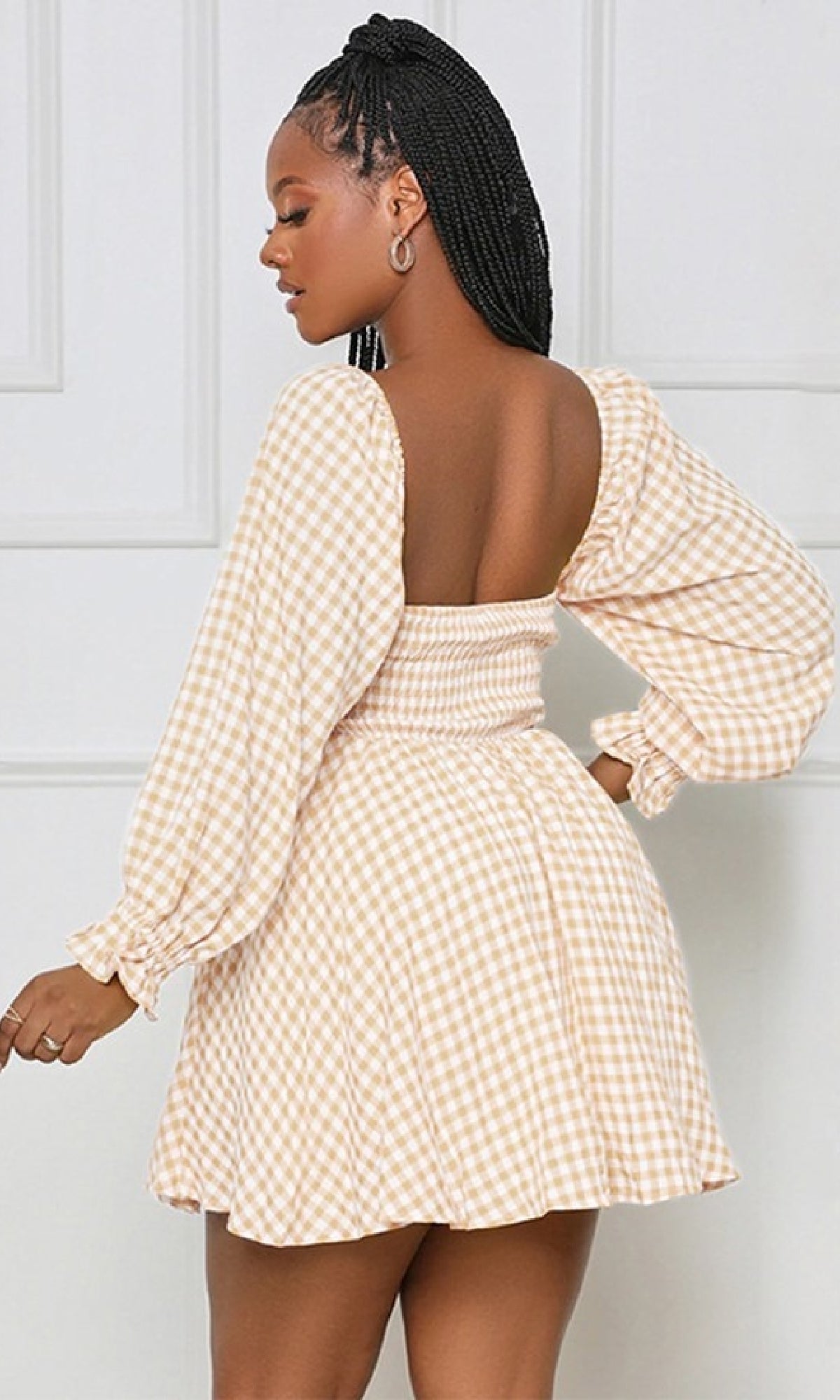 A woman stands sideways in front of a white wall wearing the Long-Sleeve Short Casual Gingham Print Dress, featuring a beige and white classic gingham pattern, open back, and effortless style.