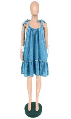 A mannequin displays the Short Casual Smock Dress with Shoulder Ties in blue, featuring a sleeveless design, ruffled hem, shoulder tie straps, and pockets, standing on a round black base.