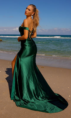 A woman wearing the Long Metallic Off-Shoulder Long Prom Dress BZ9030, featuring a corset-style bodice, stands on a sandy beach near the ocean, gazing over her shoulder.