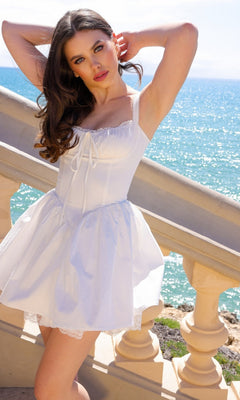 Wearing the Velvi Short White Mini Grad Party Dress Brandi, a woman poses on a seaside staircase with bright sunlight and the blue ocean in the background.