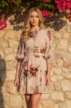 A woman wearing the Blush Floral Printed Short Casual Dress: Brooke stands in front of a stone wall with blooming pink flowers above her.