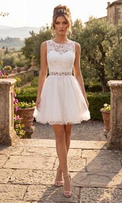 Wearing the "Short Homecoming Dress: Chicas C1107" in white, a woman stands outdoors on a stone path, surrounded by greenery and potted plants. The dress is short and sleeveless, featuring a delicate lace bodice with a fit-and-flare silhouette, complemented by heels.