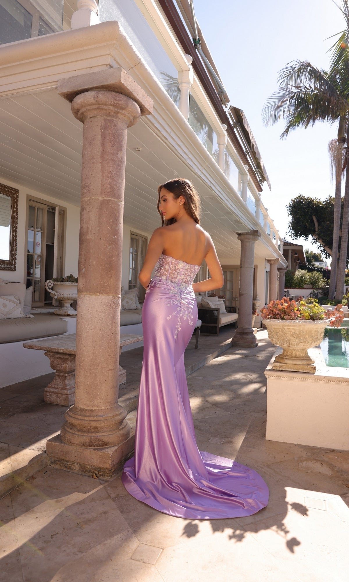 A woman wearing the Strapless Sweetheart Long Prom Dress C1346 with a sheer corset bodice stands outdoors near a column, with a pool, palm tree, and large house in the background.
