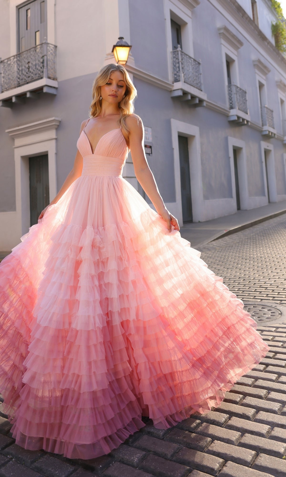 A woman in the Nox Anabel Ruffled Ombre Prom Ball Gown C1420 stands on a cobblestone street beside a light blue building in daylight.