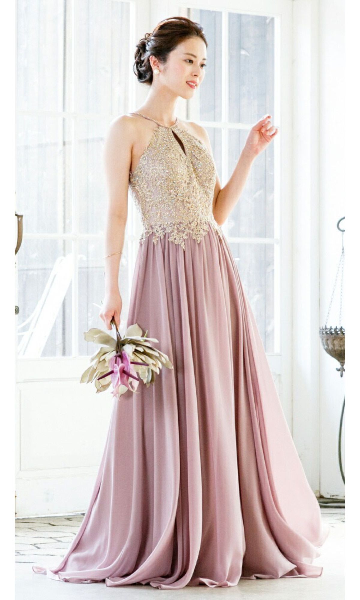 A woman indoors holds a bouquet, wearing the Chicas C1521 High-Neck Long A-Line Prom Dress—sleeveless, floor-length, in mauve with an embellished bodice and golden lace embroidery.