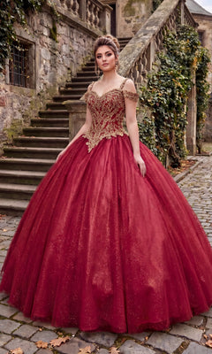 A person stands outdoors on a cobblestone path, wearing the Quinceanera Dress: Chicas C1535 in burgundy, featuring an ornate, embellished bodice with gold detailing, in front of old stone steps and ivy-covered walls.