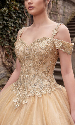 A woman wearing the Chicas C1535 Quinceanera Dress, an ornate gold off-the-shoulder ball gown with an embellished bodice featuring intricate embroidery and beadwork, stands outdoors near stone steps and greenery.