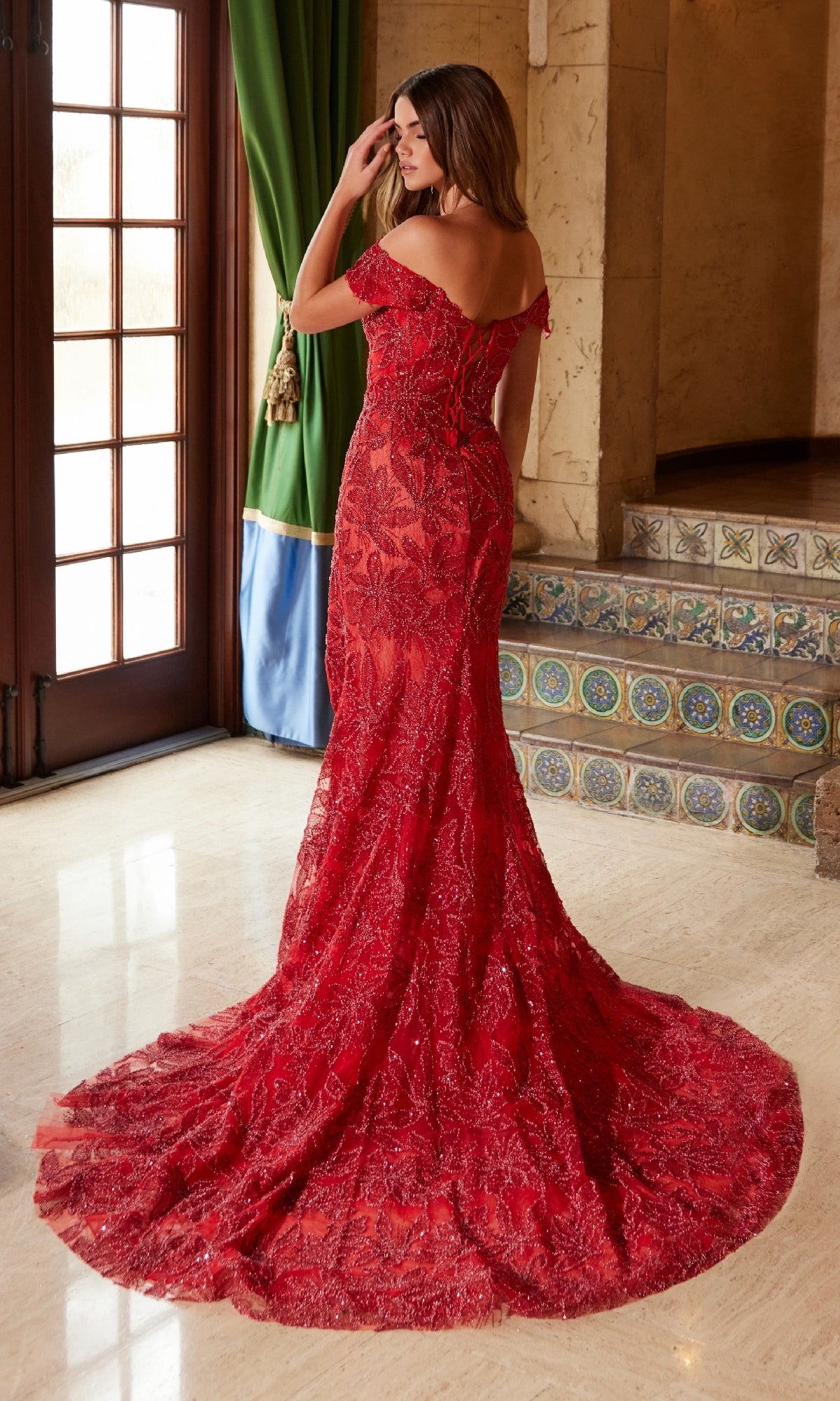 A woman wearing the Nox Anabel C1600 Lace-Up Long Beaded Prom Dress stands indoors near a window with green curtains and decorative tiled stairs.