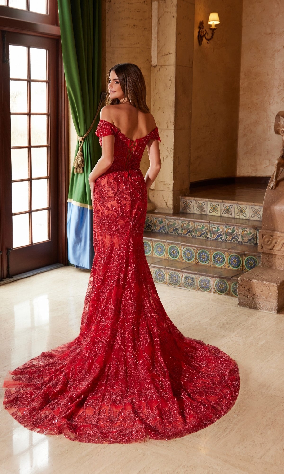A woman stands indoors near tiled steps and green curtains, wearing the Nox Anabel C1600 Lace-Up Long Beaded Prom Dress in red, featuring an off-the-shoulder lace design and a long train, facing away from the camera.