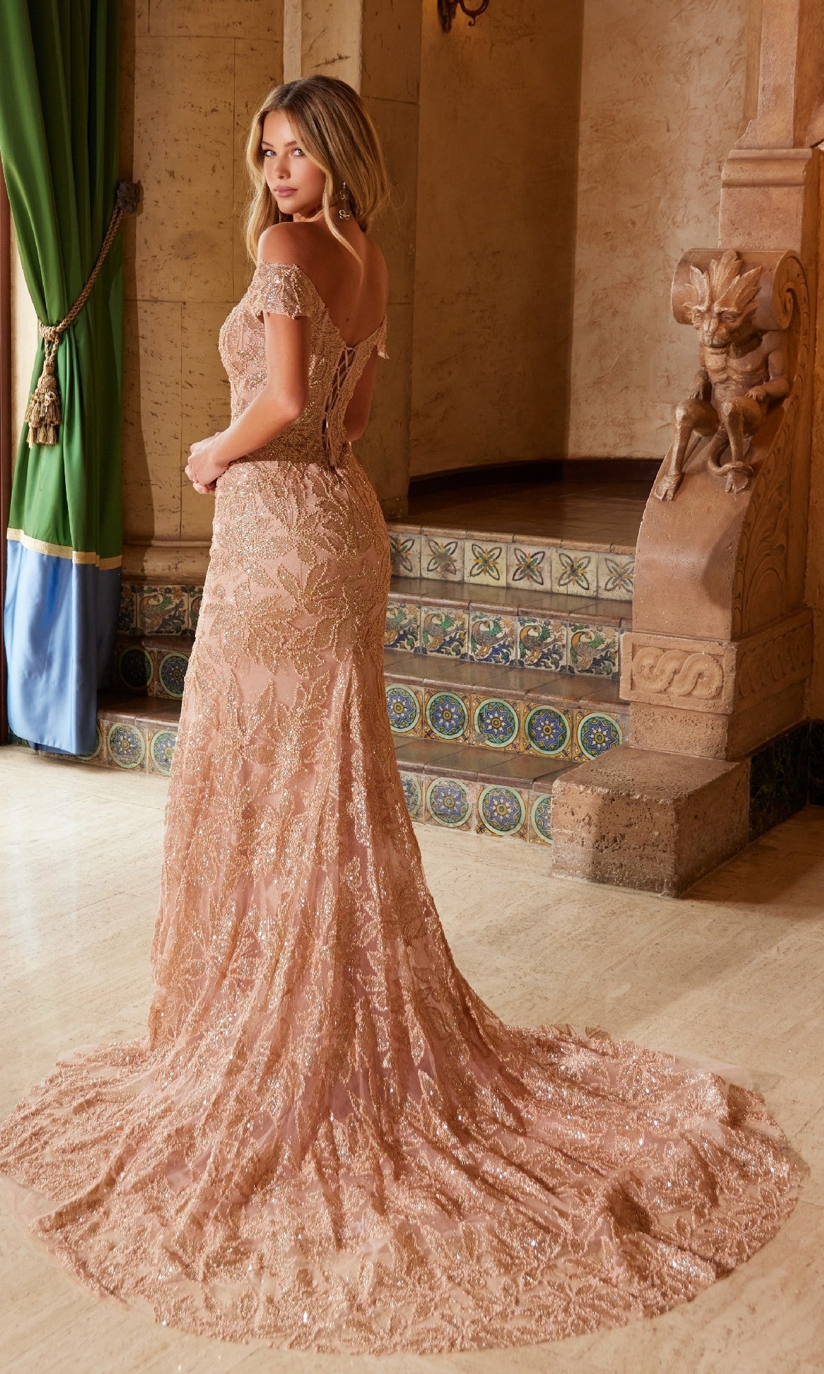 A woman in the Nox Anabel C1600 Lace-Up Long Beaded Prom Dress stands indoors by ornate tiled stairs and a green curtain, wearing a blush floor-length, off-the-shoulder gown with a long train, looking over her shoulder.