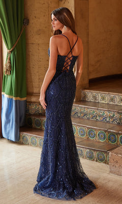 A woman stands indoors near tiled steps and green curtains wearing the Nox Anabel C1616 V-Neck Long Beaded Prom Dress in navy blue, featuring a floor-length design and lace-up back.