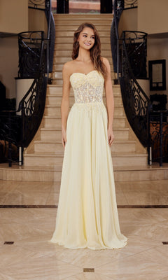A woman stands on marble flooring by a staircase, wearing the Nox Anabel C1617 strapless sweetheart prom dress in lemon with a sheer embroidered bodice featuring floral details and a flowing skirt.