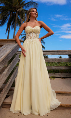 A woman in the Nox Anabel C1617 Strapless Sweetheart Prom Dress with a lace bodice and floor-length light yellow skirt stands on a wooden walkway at the beach, framed by palm trees and a blue sky.
