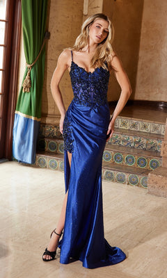 A woman in the Nox Anabel C1625 Beaded-Corset Long Prom Dress, featuring a blue hue, black floral embellishments, and a thigh-high slit, stands indoors near tiled steps.
