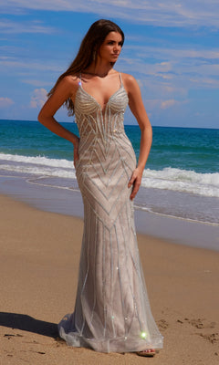 Wearing the Long Glitter-Lace Prom Dress Nox Anabel C1632—a sleeveless, floor-length gown with geometric beading and a lace-up open back—a woman stands on a sandy beach by the ocean under a partly cloudy sky.
