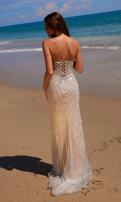 Wearing the Nox Anabel C1632 Long Glitter-Lace Prom Dress, a woman stands on a sandy beach facing the ocean, the gown’s lace-up open back and shimmering fabric catching the sunlight as waves roll in.