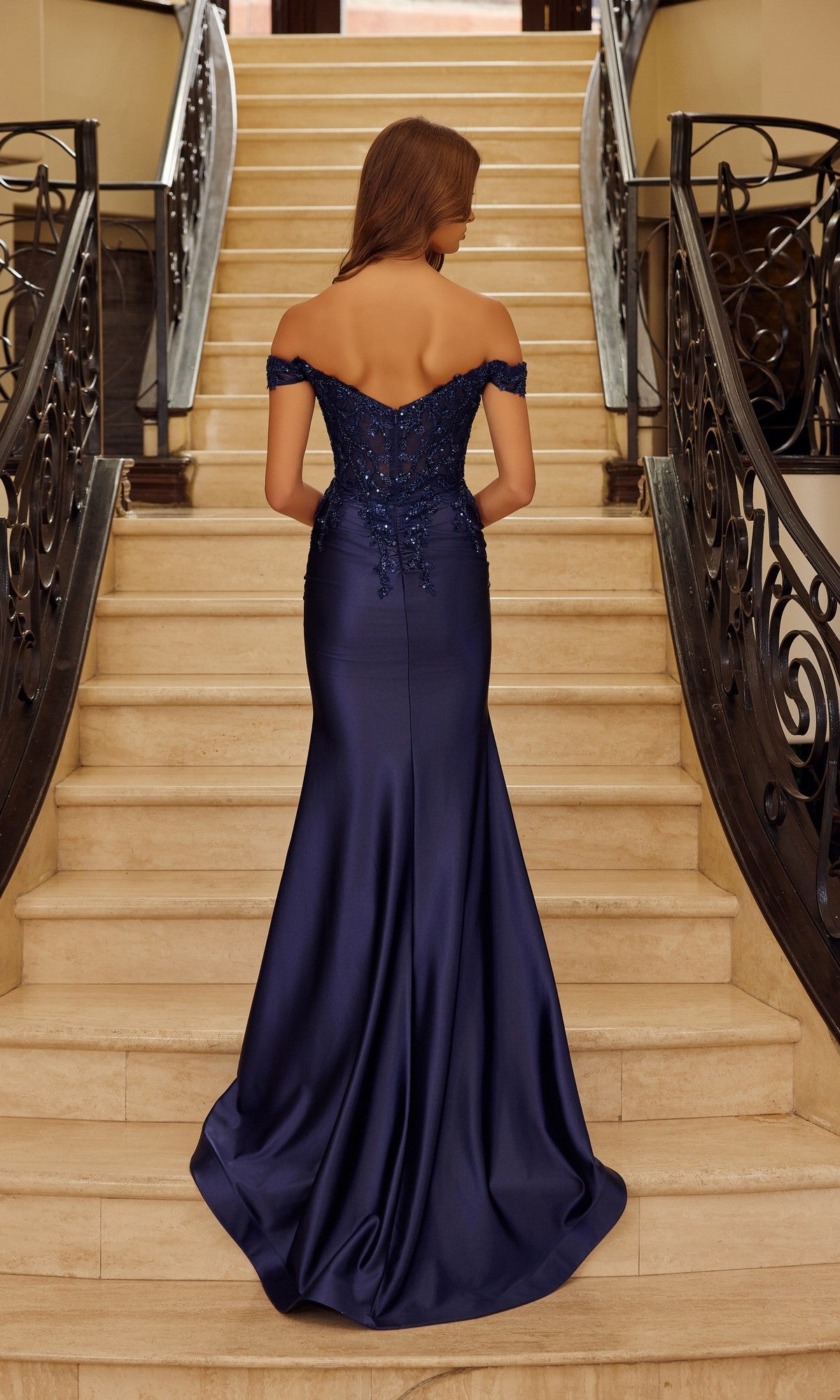 A woman in the Nox Anabel C1647 off-the-shoulder long prom dress, featuring navy blue mermaid style and delicate beaded embroidery, stands on a staircase facing away from the camera.