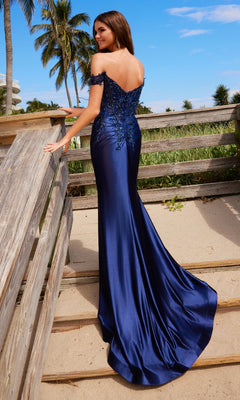 A woman in the Nox Anabel C1647 Off-the-Shoulder Long Prom Dress with a mermaid silhouette and long train stands on a sandy beach near a wooden fence, with palm trees in the background.