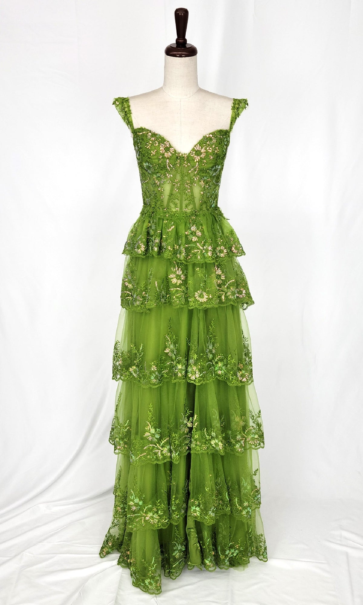 The Chicas C2433 Tiered Long Ruffle Prom Dress is a floor-length, sleeveless green gown with floral embroidery and layered ruffles, displayed on a dress form against a white background.