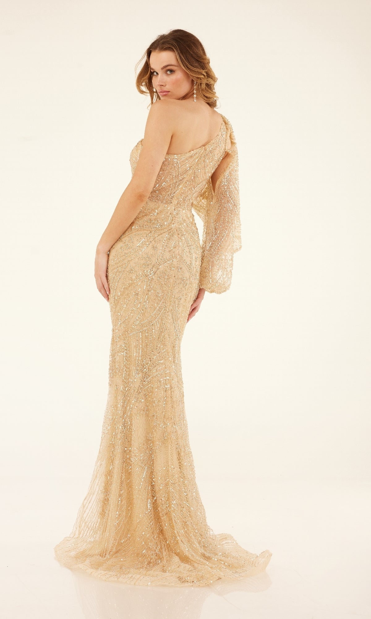 A woman poses in the Cecilia Couture C273A, a floor-length gold prom dress with one-shoulder design and sparkling sequin embellishments, standing and looking over her shoulder against a plain background.