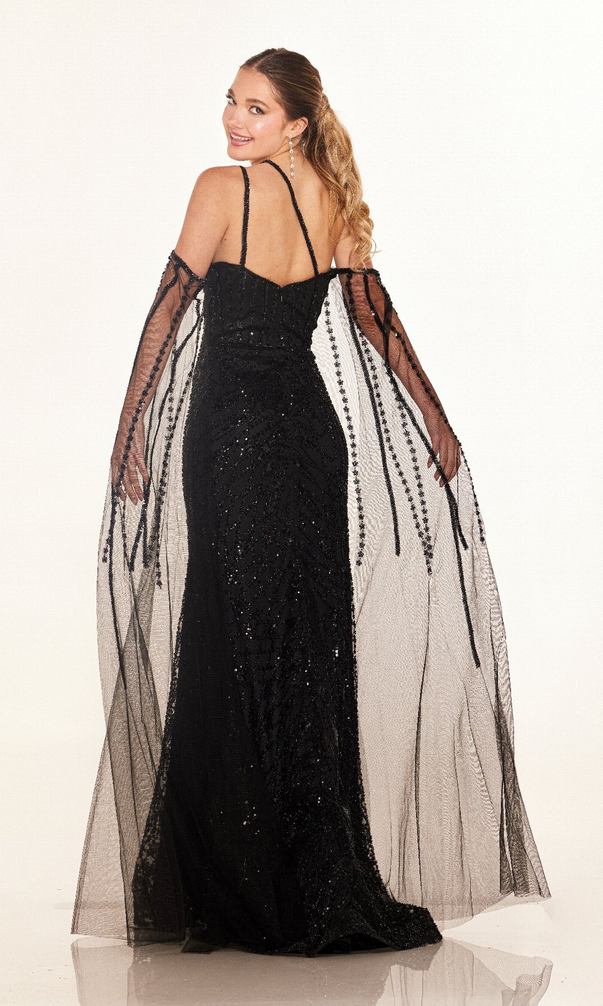 Woman wearing the Cecilia Couture C280, a sparkly long formal dress with a high leg slit and sheer black shawl, stands against a white background and looks back over her shoulder.
