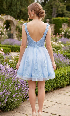 A woman with blonde hair in a low bun wears the Chicas C311 short homecoming dress in pastel blue with floral embroidery, standing on a garden path facing away from the camera.