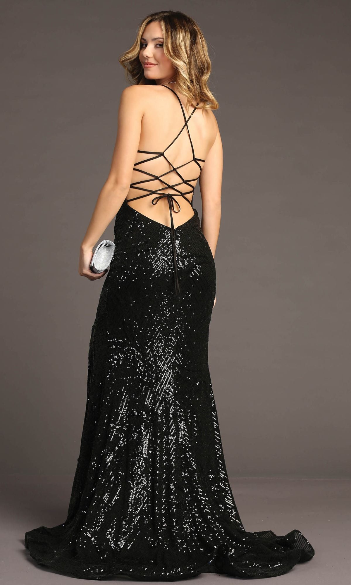 A woman in the Chicas C316 Lace-Up Long Sequin Prom Dress, featuring a black floor-length design and open back, holds a silver clutch against a plain background.