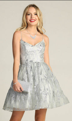 A woman with long blonde hair, red lipstick, and a silver necklace smiles at the camera while wearing the Lace-Up Glitter-Print Short Homecoming Dress C317 and holding a matching clutch.