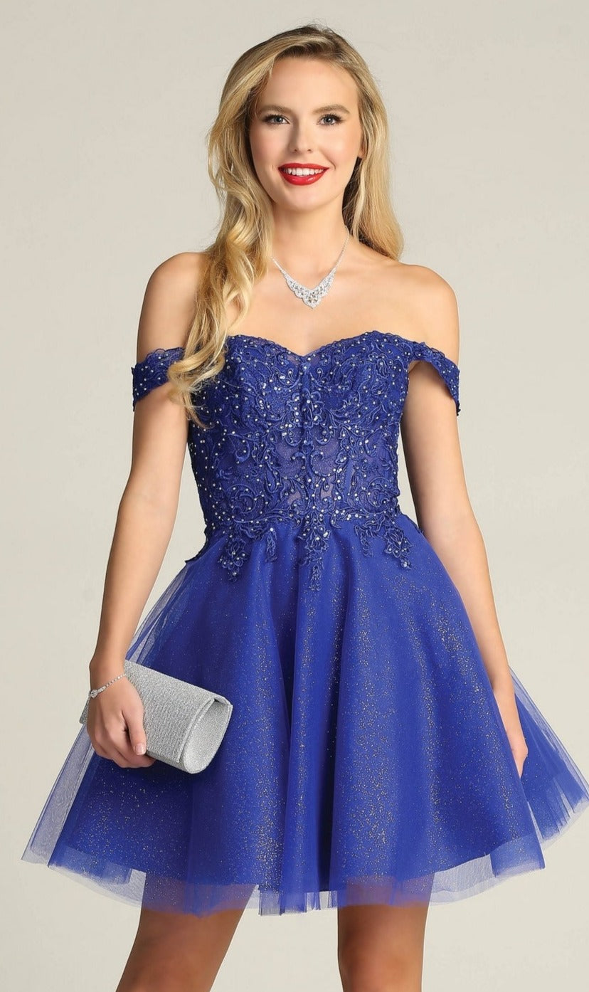 A woman wears the Off-Shoulder Short Glitter Homecoming Dress C318 in royal blue, featuring sequin details. She accessorizes with a silver clutch, necklace, and bracelet for a sparkling party look.