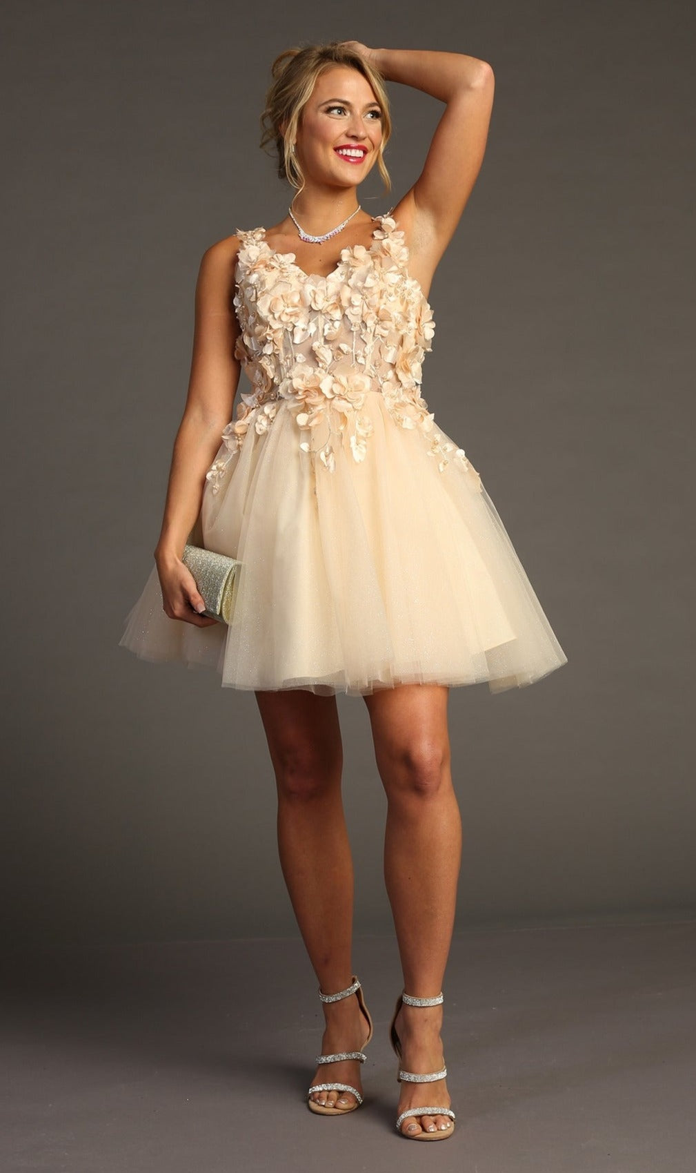 A woman models the Floral-Bodice Short Babydoll Homecoming Dress C321—a sleeveless, knee-length beige dress with tulle and appliqué—holding a clutch with one arm raised against a plain backdrop.
