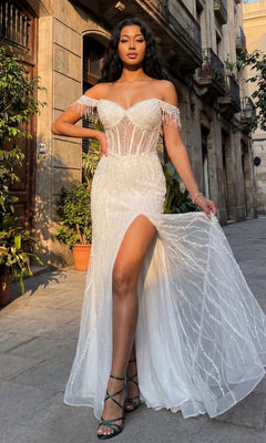 A woman poses outdoors on a city street wearing the Chicas C331 Off-Shoulder Long Sequin Prom Dress, featuring a white beaded design, high slit, and transparent corset bodice.