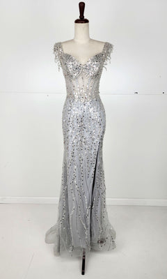 The Off-Shoulder Long Sequin Prom Dress: Chicas C331 is shown as a stunning silver gown with a fitted bodice, sheer panels, and sparkling embellishments, displayed on a dress form against a plain white background.