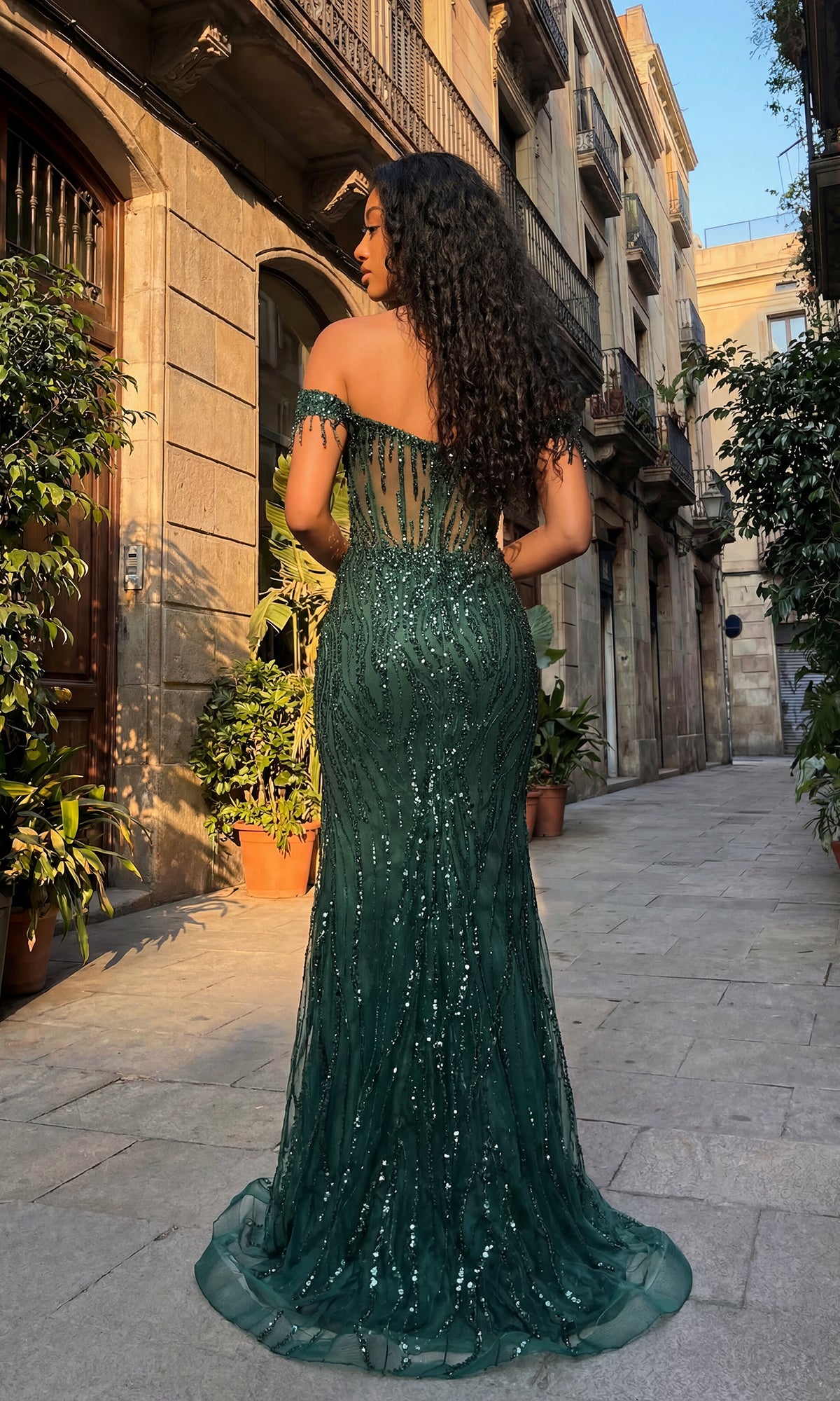 A person in the Off-Shoulder Long Sequin Prom Dress: Chicas C331 stands on a cobblestone street lined with plants and old buildings, facing away from the camera.