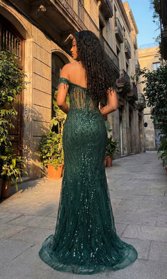 A person in the Off-Shoulder Long Sequin Prom Dress: Chicas C331 stands on a cobblestone street lined with plants and old buildings, facing away from the camera.
