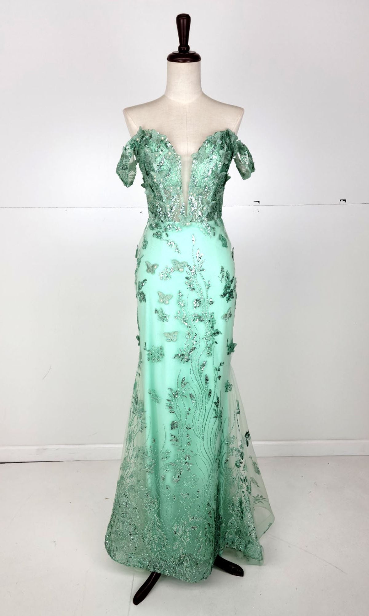 The Off-Shoulder Long Butterfly Prom Dress: Chicas C332 is shown on a dress form, featuring mint green color, a deep V-neckline, sparkling floral embellishments, and a fitted mermaid silhouette.