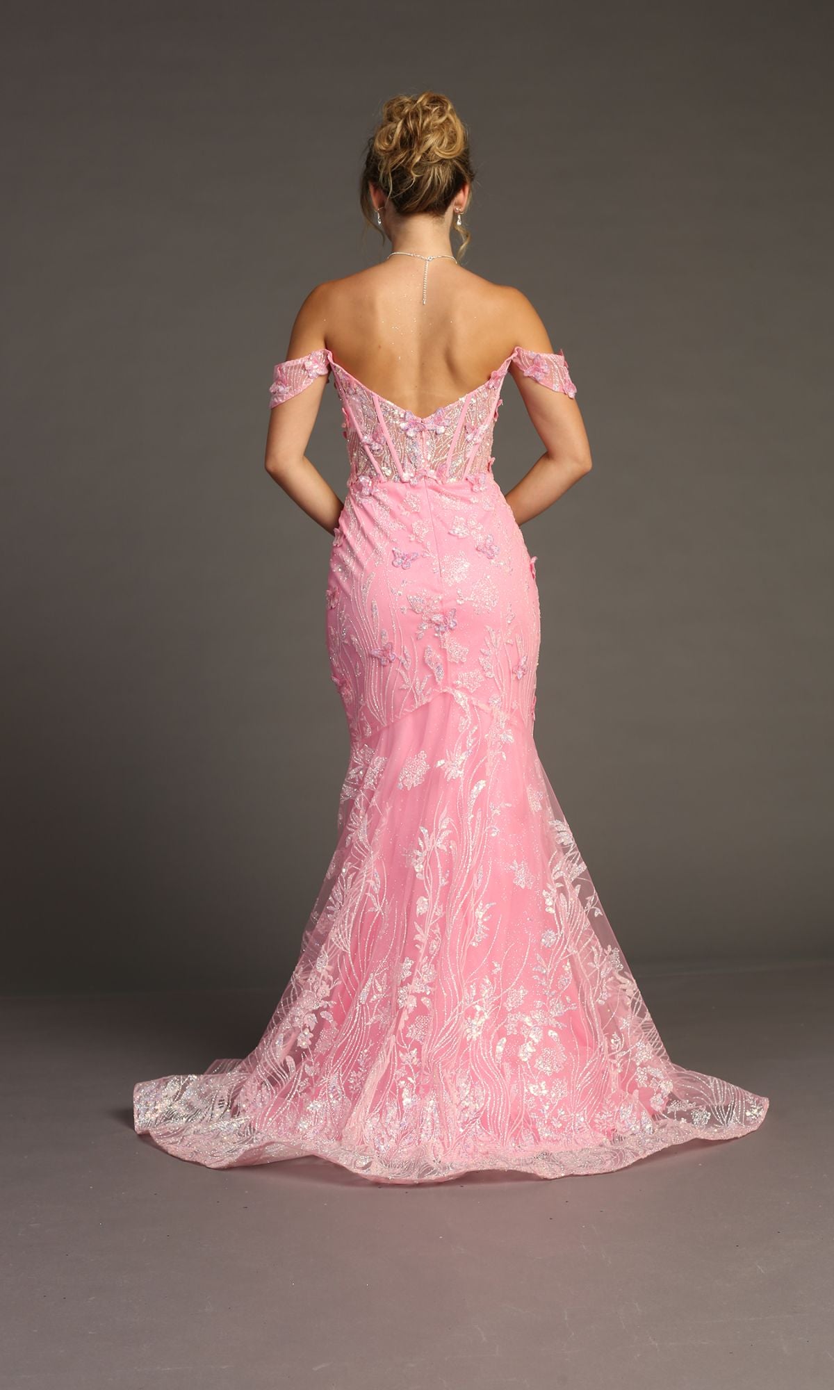 A woman faces away in the Off-Shoulder Long Butterfly Prom Dress: Chicas C332—a floor-length pink gown with floral embroidery and flared hem—posing against a plain gray background.