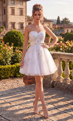 A woman wearing the Chicas C3350 Short Homecoming Dress in white, featuring a strapless fit-and-flare silhouette with beautiful embellishments, stands outdoors on a stone terrace with a lush garden and large house in the background. The elegant dress highlights her figure, adding a refined touch to the picturesque scene.