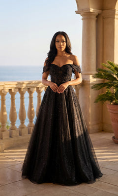 A woman wears the Chicas C336 long prom dress—black, off-the-shoulder, floor-length with an adjustable lace-up back—while standing on a sunlit balcony overlooking the ocean and stone pillars.