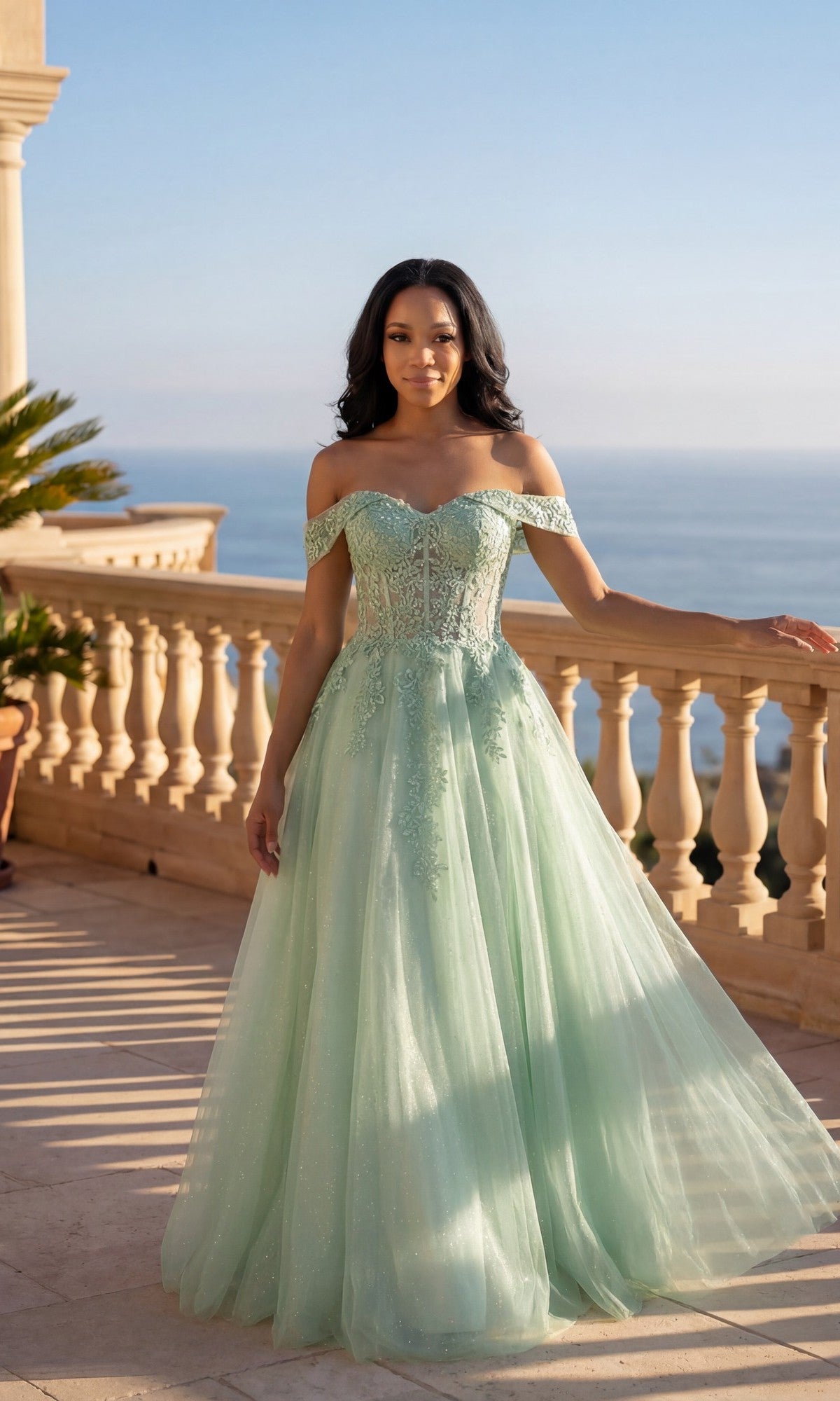 A woman in a sage Long Prom Dress: Chicas C336 with floral lace embroidery stands on a sunlit terrace, the sea view behind her.
