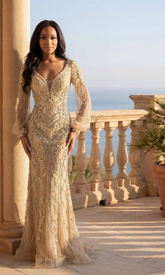 A woman stands on a stone balcony overlooking the sea, wearing the Chicas C339 long formal dress in champagne, featuring gold embroidery, lace appliqué, and sheer sleeves.
