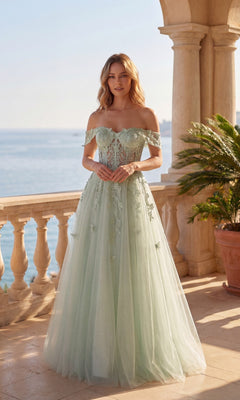 A woman in the Chicas C350 Long Prom Dress—sage, off-the-shoulder glitter tulle with floral accents and a corset bodice—stands on a stone terrace by the sea, beside a potted plant.