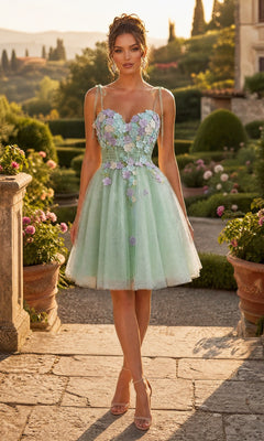 A woman wearing the Short Homecoming Dress: Chicas C354 in sage, featuring 3D floral appliqués and an a-line tulle short skirt, paired with nude heels, stands in a sunlit garden surrounded by flowers and stone architecture.